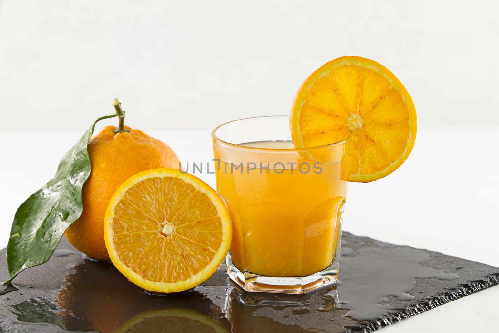 An inviting glass full of orange juice with orange slice on the rim, a half orange and a whole one with leaf on a square wet slate plate on white background
