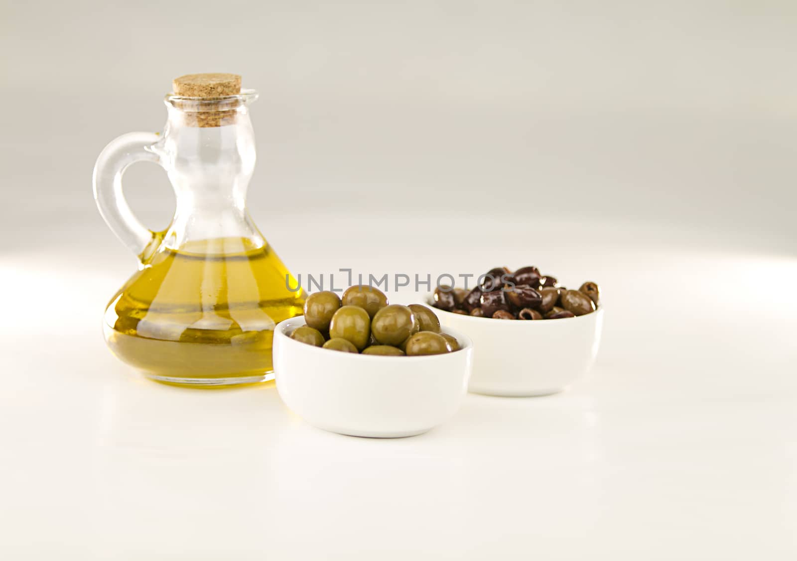 Close-up of a flask with extra virgin olive oil and two small cups with different varieties of olives on white background