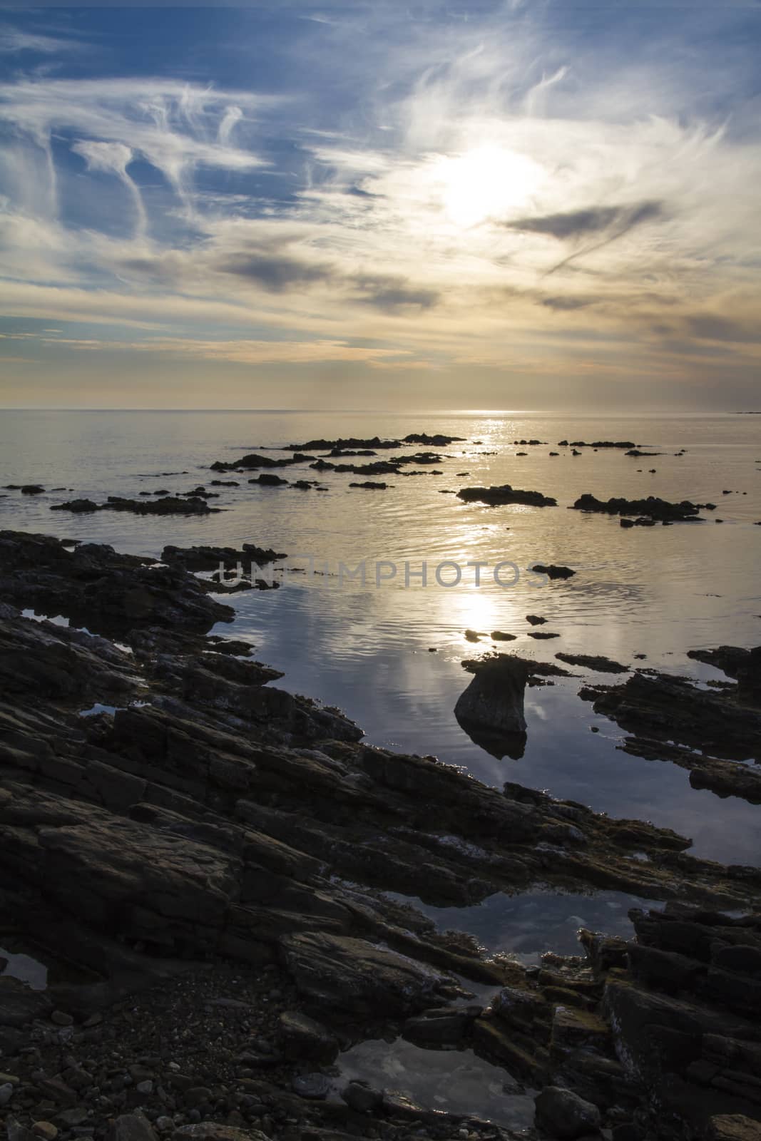 Blue sky at dawn covered by strange cloud formations on a flat sea with black rocks that stand out in the reflection of the sun on the water