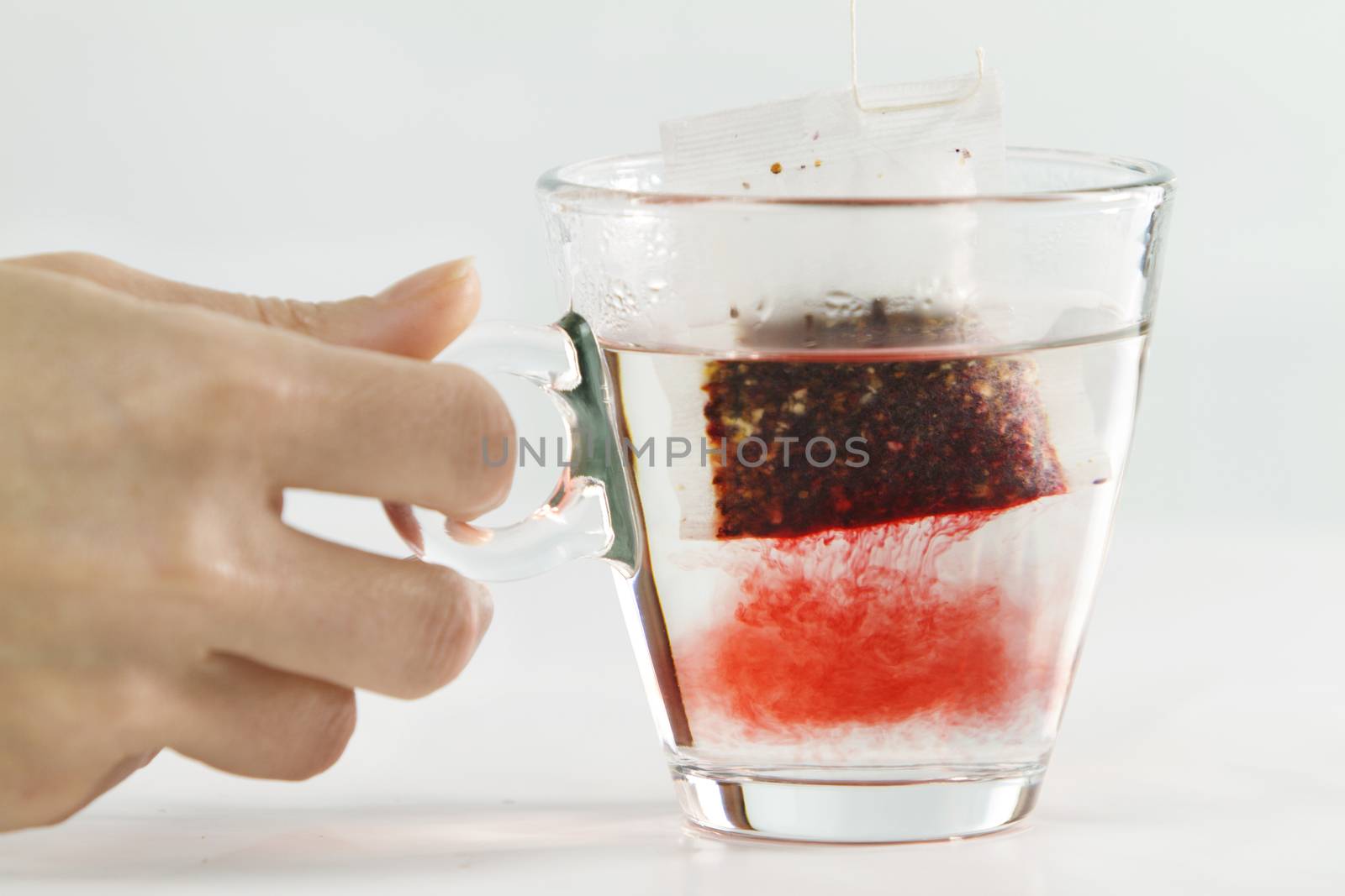 Close-up of a woman hand dipping a sachet of Hibiscus Tea in a glass cup full of water with beautiful red-colored effects in the transparency of water