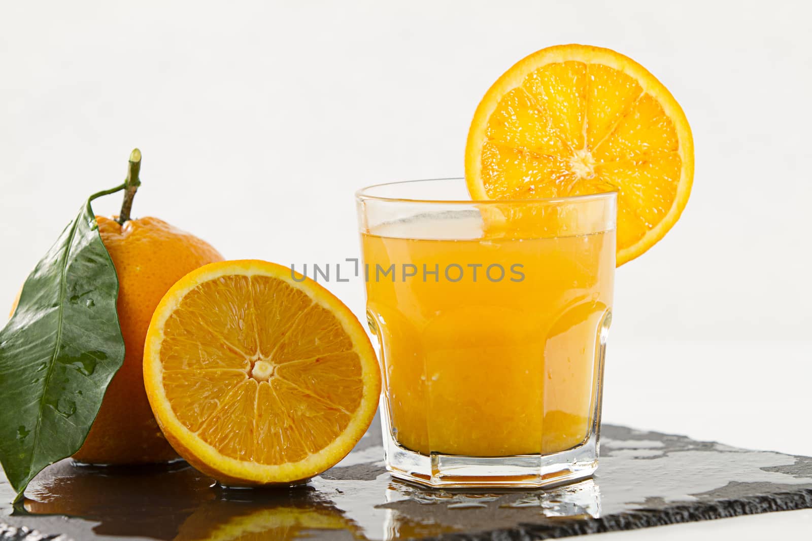 An inviting glass full of orange juice with orange slice on the rim, a half orange and a whole one with leaf on a square wet slate plate on white background