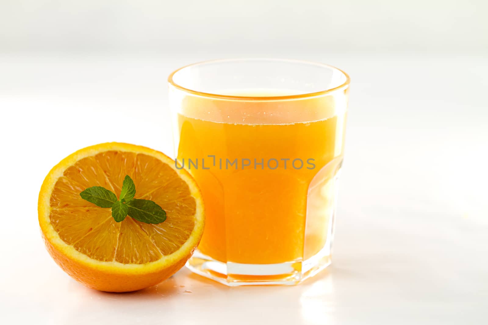 An inviting glass full of orange juice and a half one with a fresh mint leaf in its center on a white background