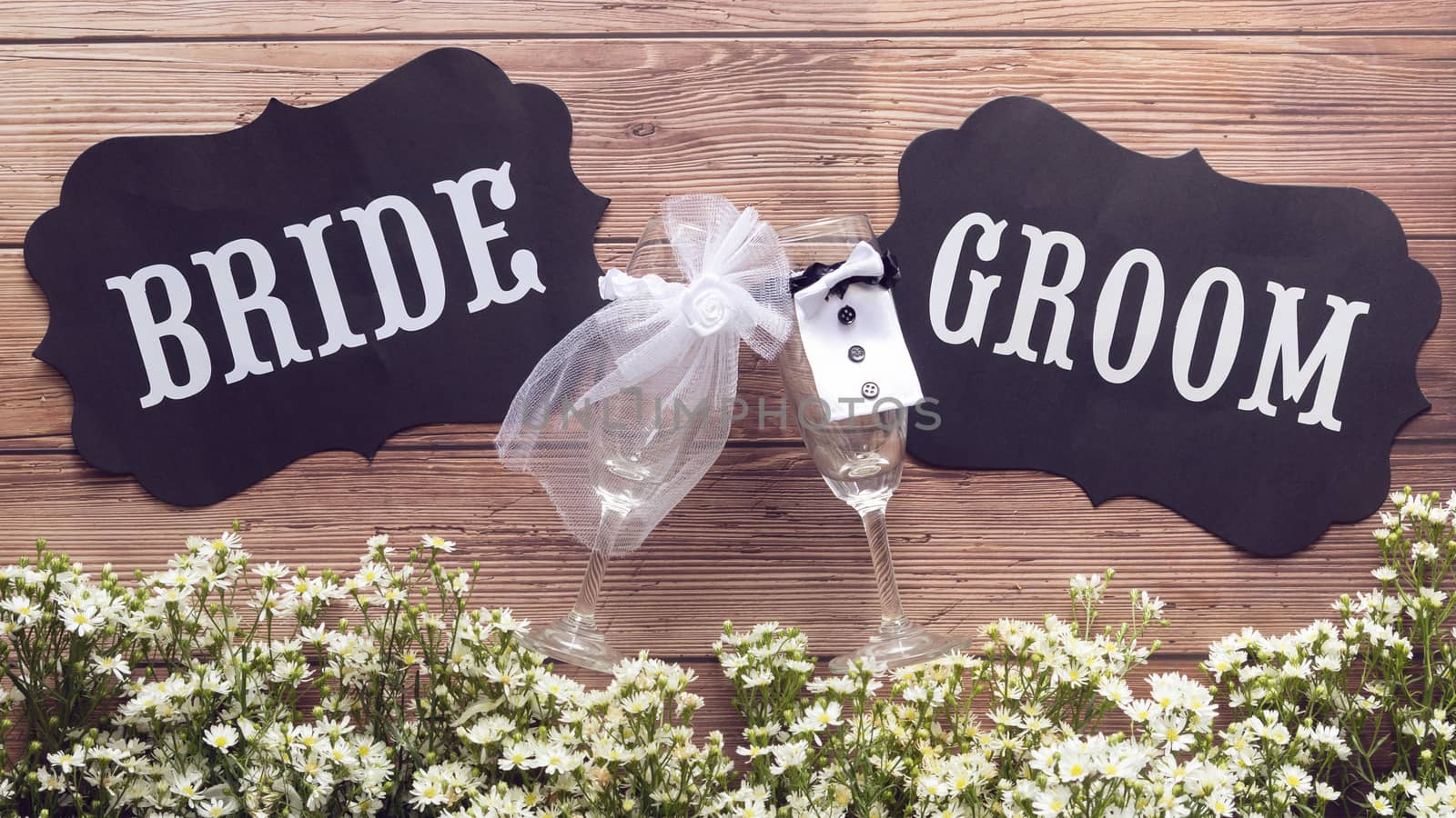 champagne glass in wedding dress with Bride and Groom text sign on wooden background decorated with tiny white flower, vintage style. wedding sign concept by asiandelight