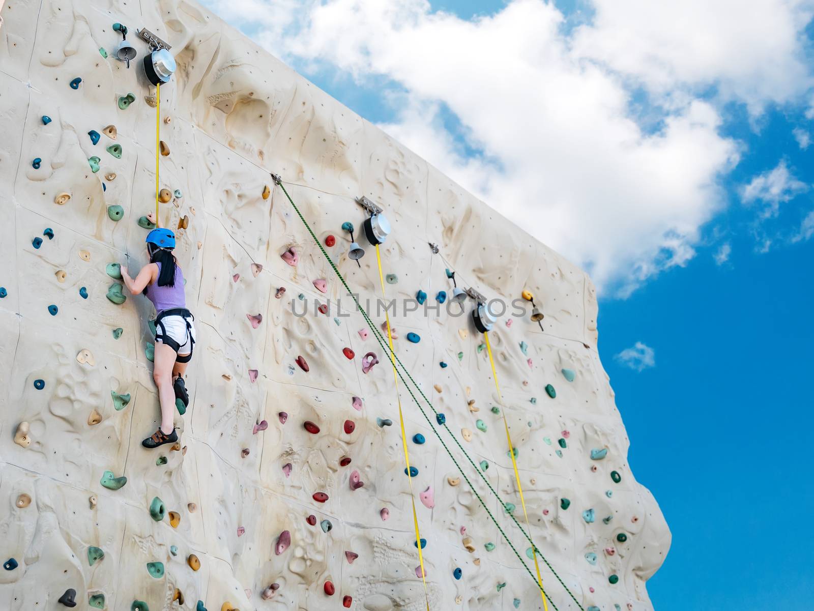 Asian athletic woman practicing rock climbing on a rock wall, outdoor activity with blue sky background, low angle view by asiandelight