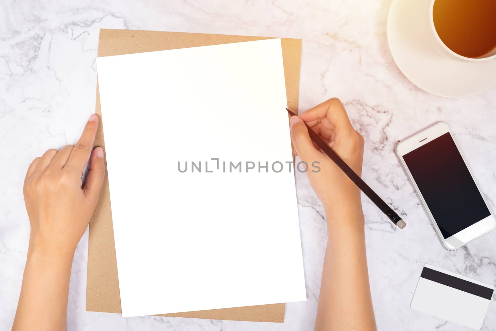flat lay of woman hand writing in a blank white paper on brown paper envelope with pencil ,mobile phone , credit card and black coffee on white marble background, top view