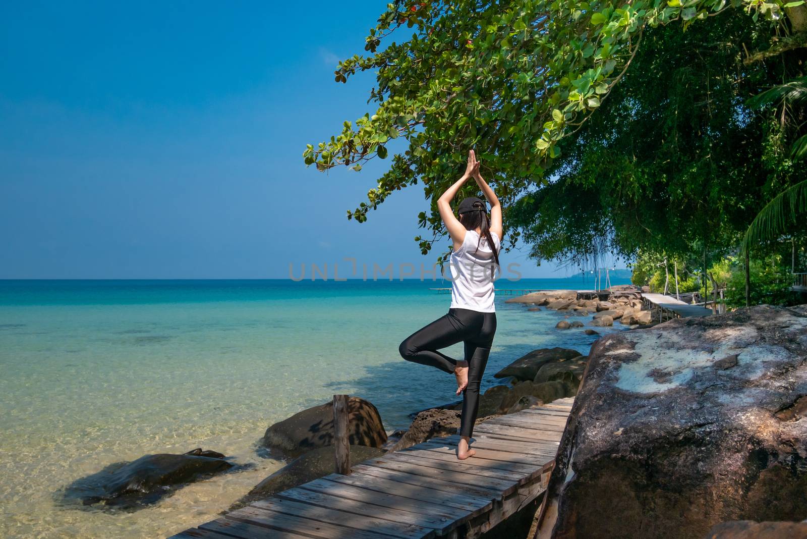 a woman standing on one leg while practicing yoga on wooden bridge over the sea during summer vacation. attractive young Asian woman standing in yoga pose by asiandelight