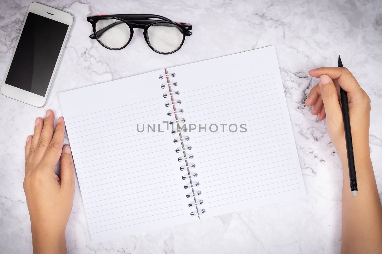 flat lay of woman hand writing in a blank white page notebook on white marble desk, top view. glasses and mobile smart phone on desk for decoration by asiandelight