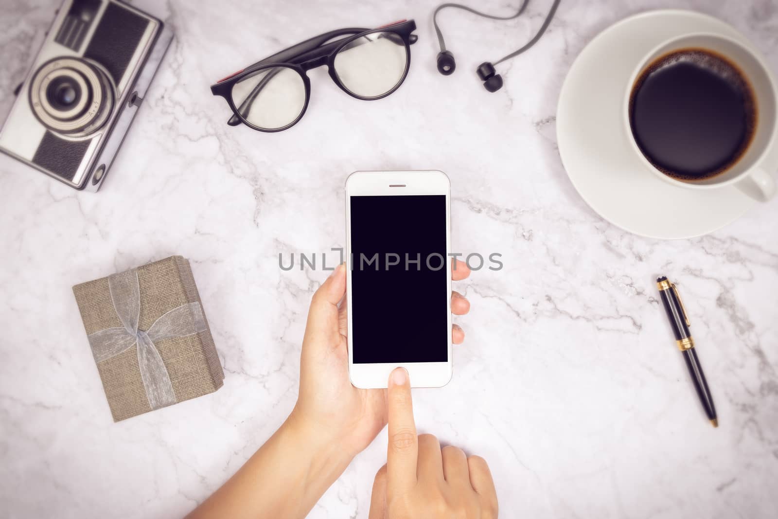 woman hand use mock up of mobile phone blank black screen with finger on touch screen with earphone,  pen, camera, glasses , cup of coffee and gift box. flat lay top view work space , duo tone