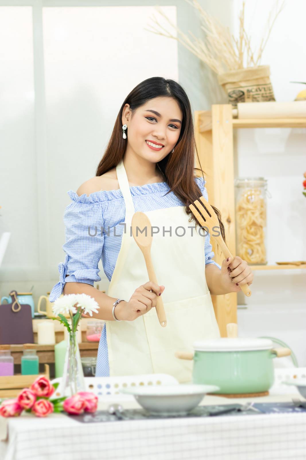 beautiful Asian woman holding wooden cutlery behind dining table with pot , dish and kitchenware in the kitchen room with smiley face by asiandelight