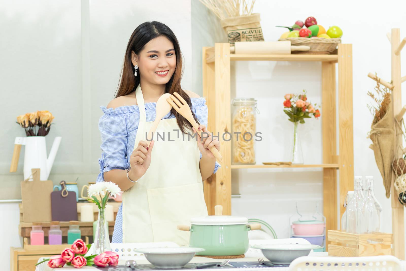 beautiful Asian woman holding wooden cutlery behind dining table with pot , dish and kitchenware in the kitchen room with smiley face by asiandelight