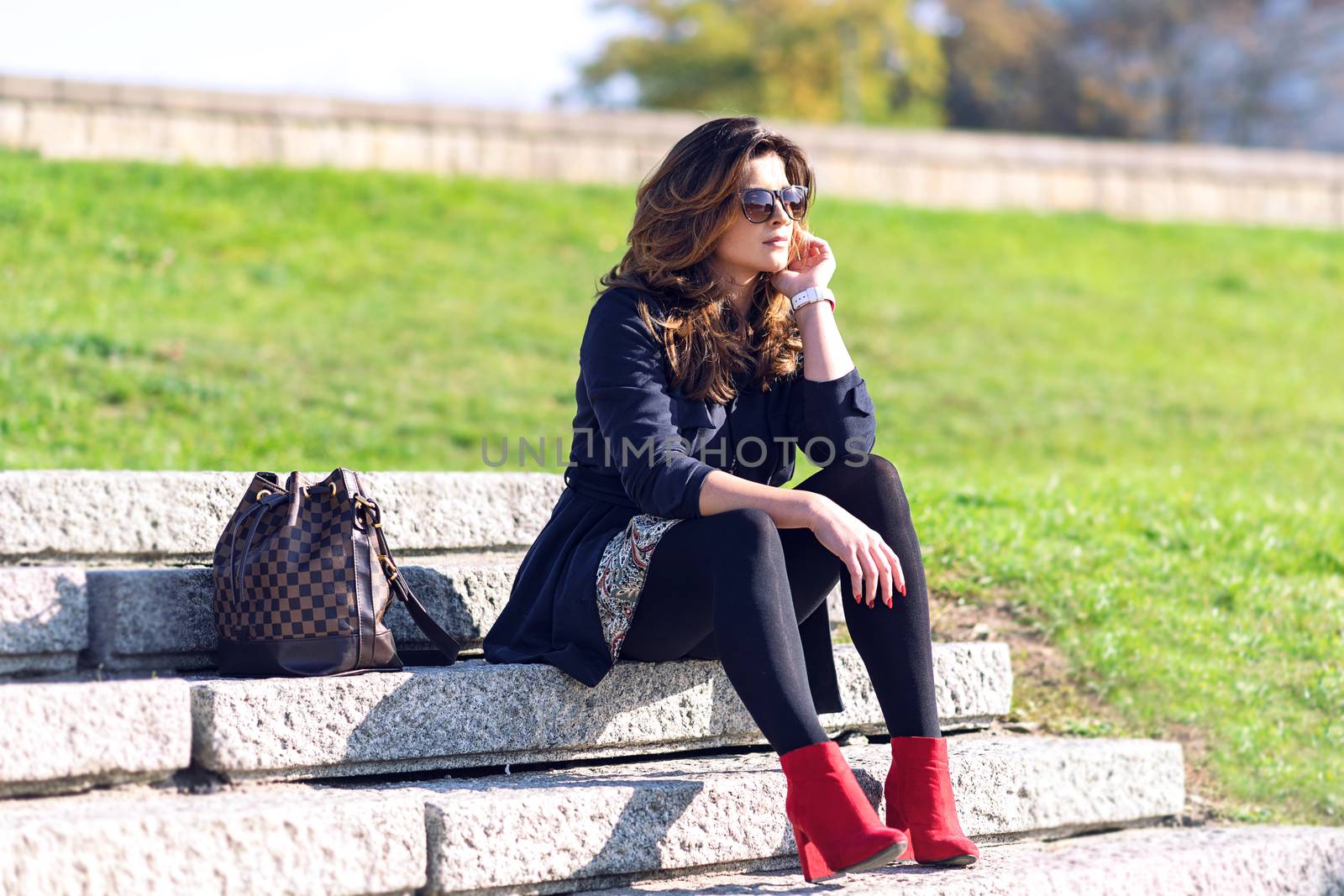 Beautiful young fashionable woman with a nice hairstyle and sunglasses sitting on the stairs and resting outdoors on a sunny day.