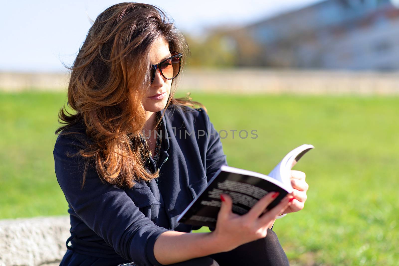 Beautiful young woman with a nice hairstyle and sunglasses sitting outdoor and reading a book on a sunny day.