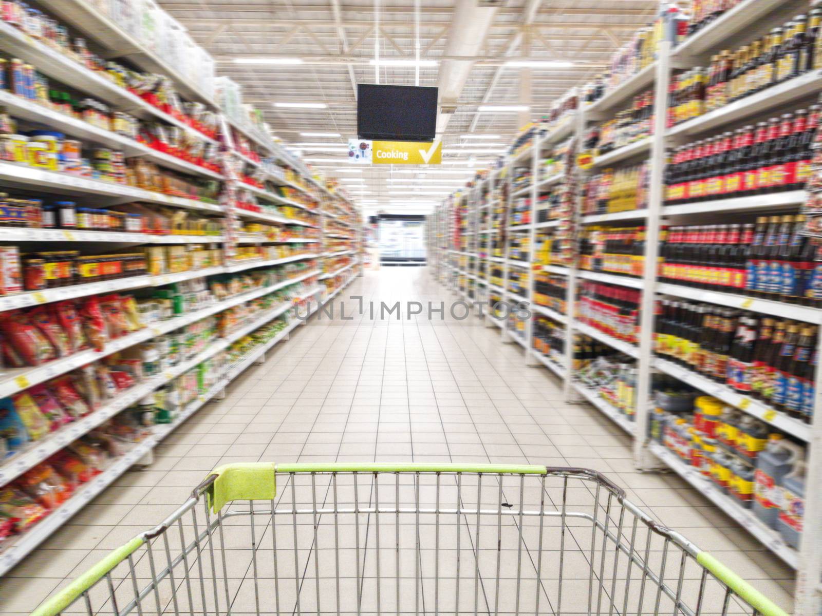 shopping cart with abstract blurred supermarket aisle with colorful goods on shelves at background, sign of product for cooking hanging from ceiling