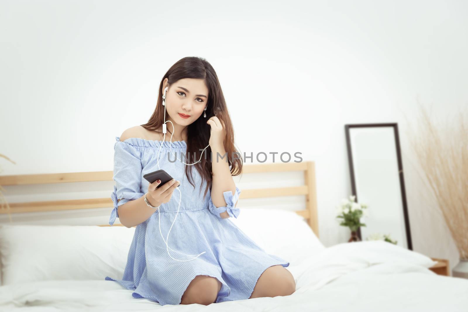 A beautiful Asian woman with headphone relaxing at home , sitting on the bed listening to music from application on mobile phone