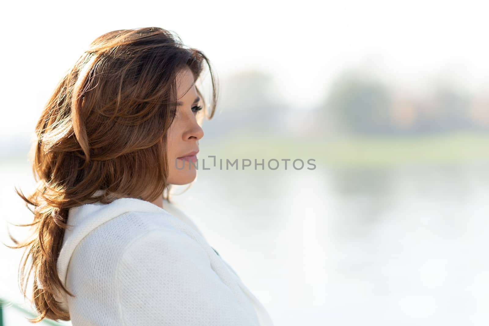 Beautiful and sensual woman is looking thoughtful at the beautiful landscape of the coast or river in the city illuminated by deep morning sunlight.