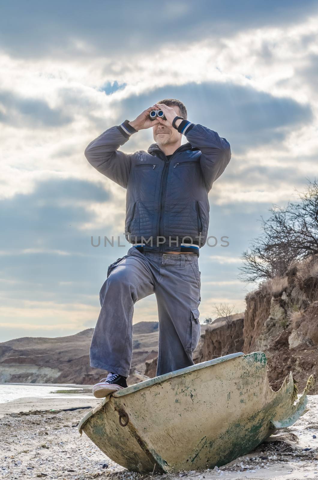 man in black stands in a broken boat on the beach and carefully looks through binoculars