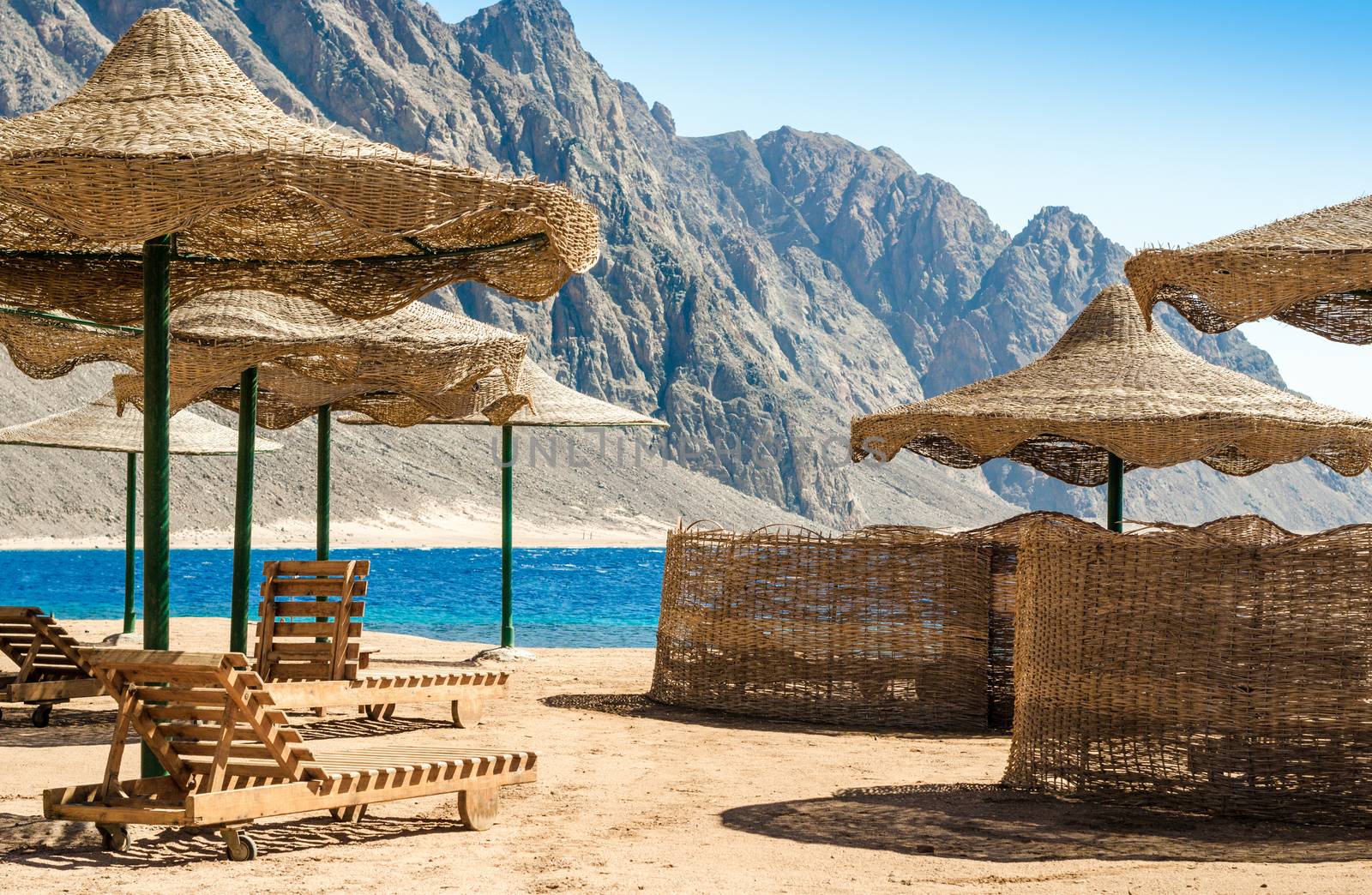 beach umbrellas and wooden lounge chairs on the sand of the beach against the backdrop of the sea and high rocky mountains