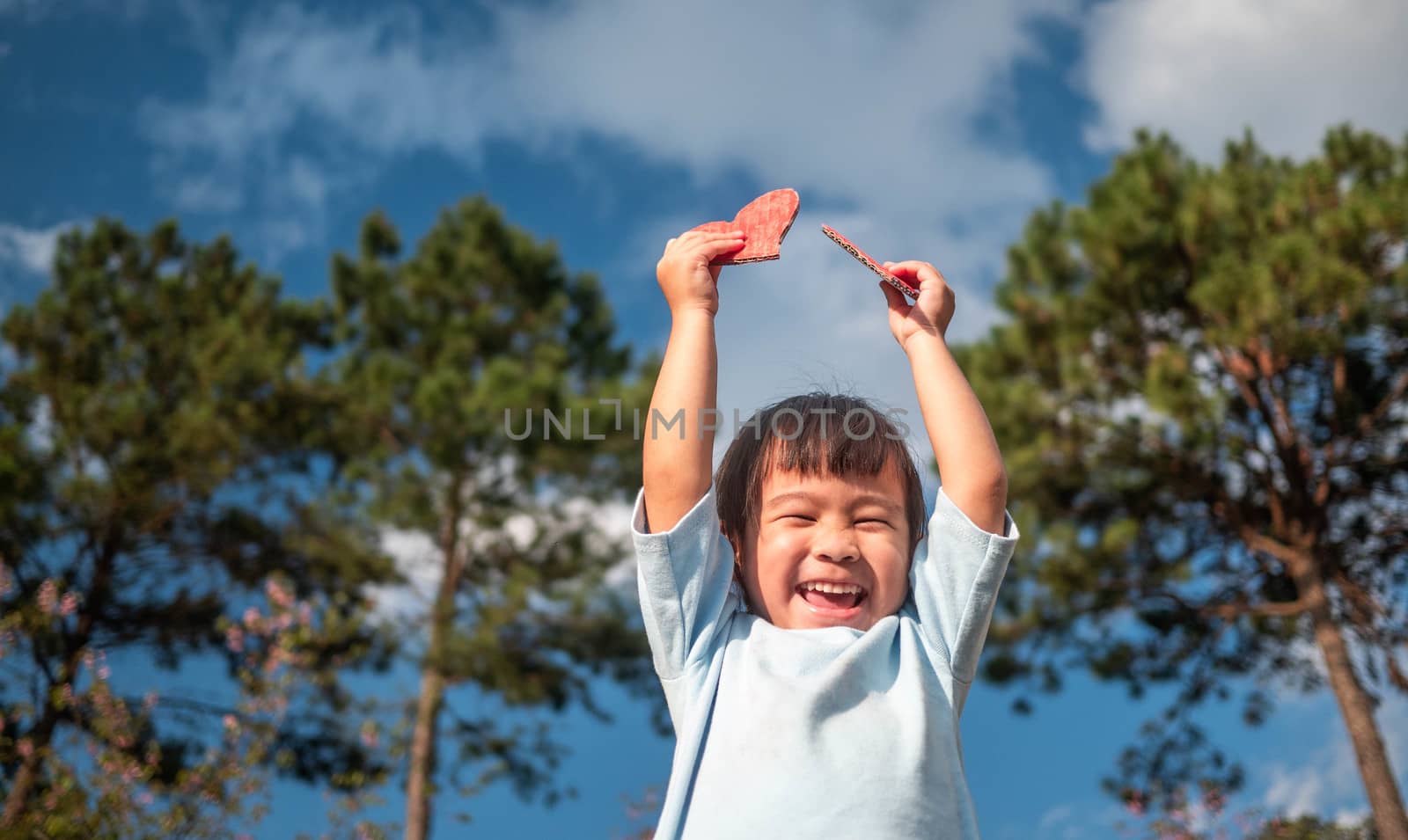 Cute little girl holding Two red heart-shaped recycled cardboard on blue sky and green tree background. Love symbol for valentines day. Concepts of Love and Romance.