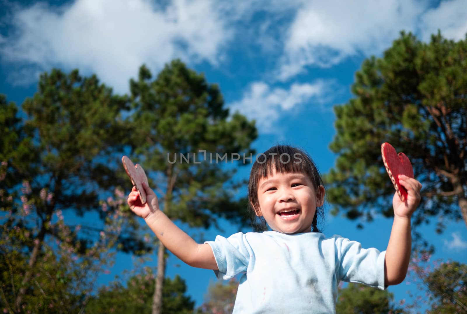 Cute little girl holding Two red heart-shaped recycled cardboard on blue sky and green tree background. Love symbol for valentines day. Concepts of Love and Romance. by TEERASAK