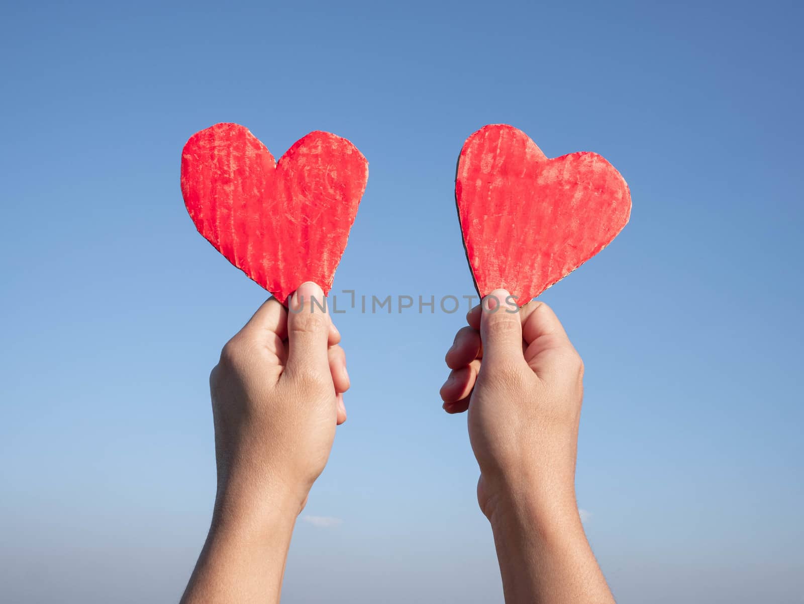 Hands holding two red heart-shaped recycled cardboard on blue sky background. Love symbol for valentines day. Concepts of Love and Romance. by TEERASAK