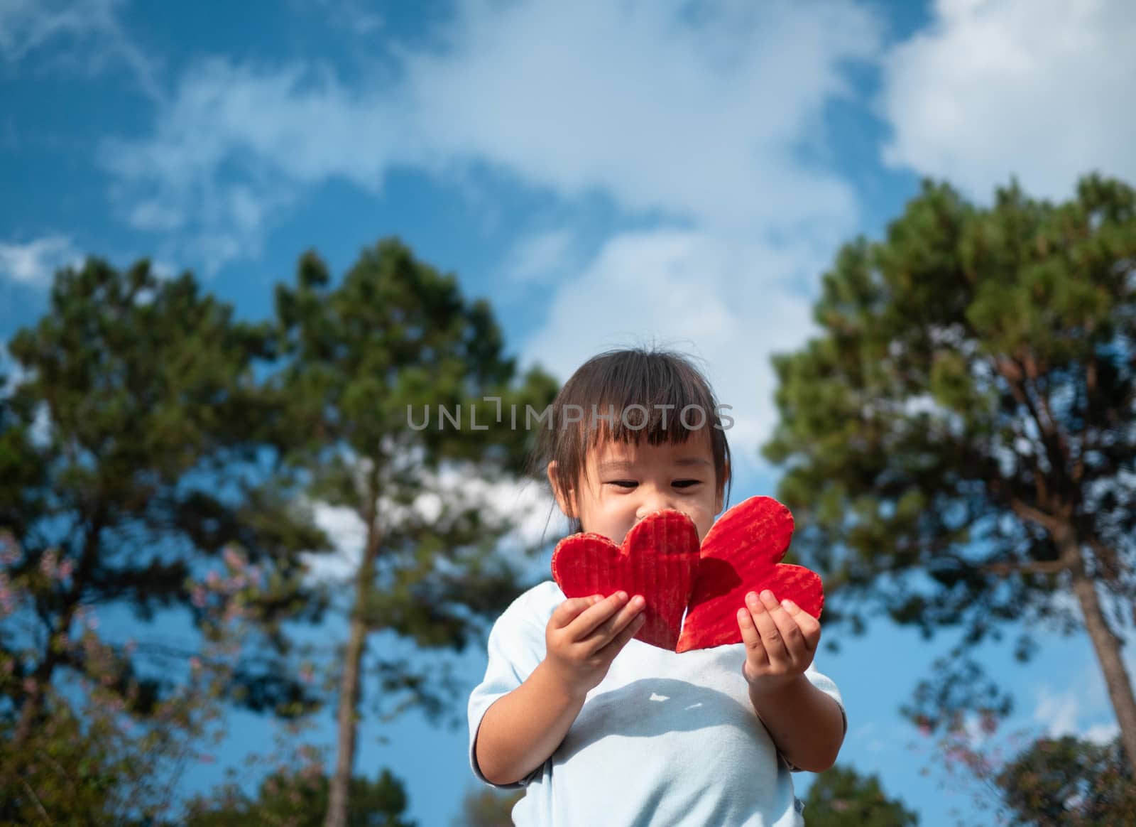 Cute little girl holding Two red heart-shaped recycled cardboard on blue sky and green tree background. Love symbol for valentines day. Concepts of Love and Romance. by TEERASAK