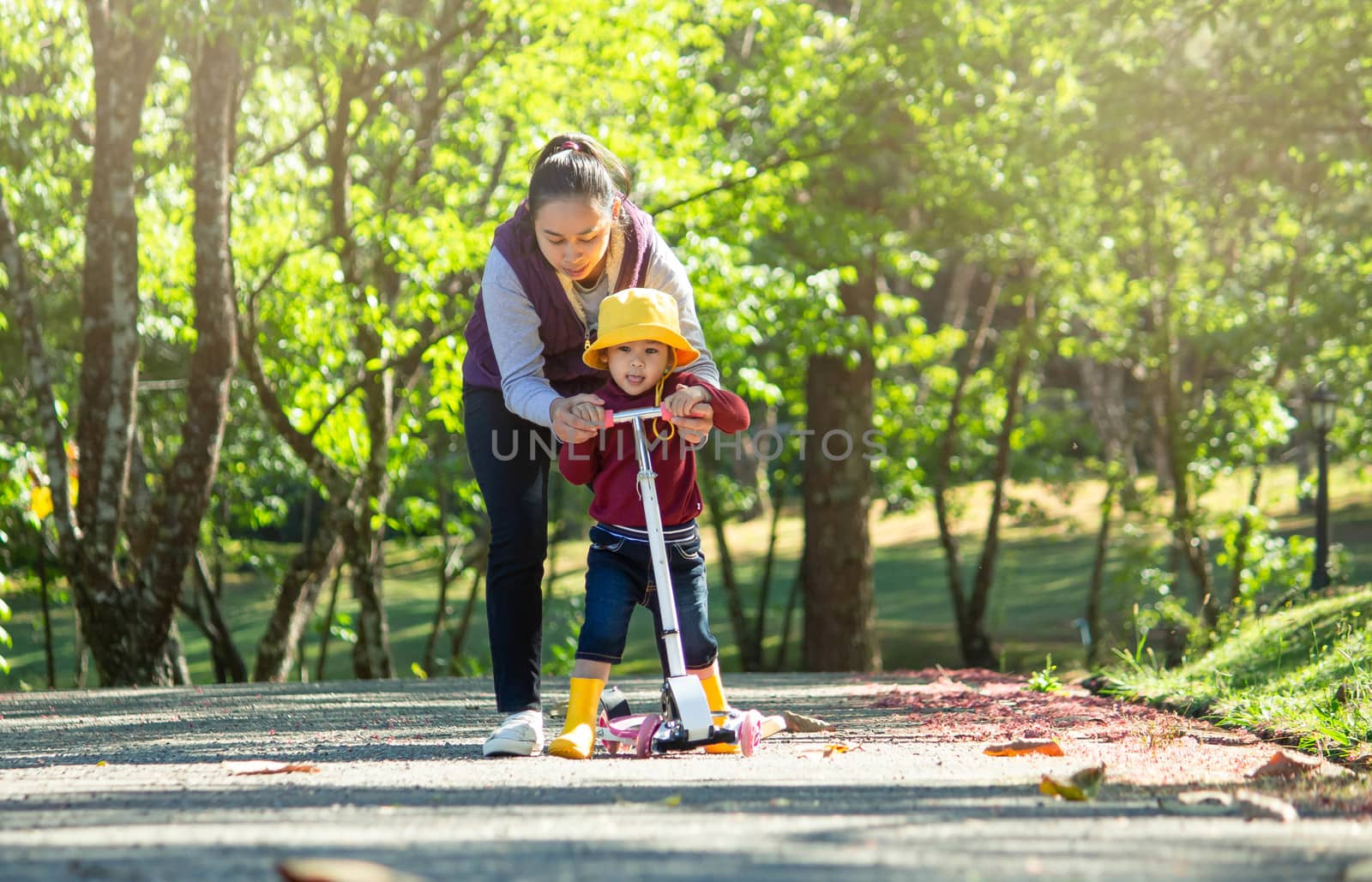 Cute little child girl having fun to riding a scooter on road with her mother in a summer park. Concept of happy family and childhood.