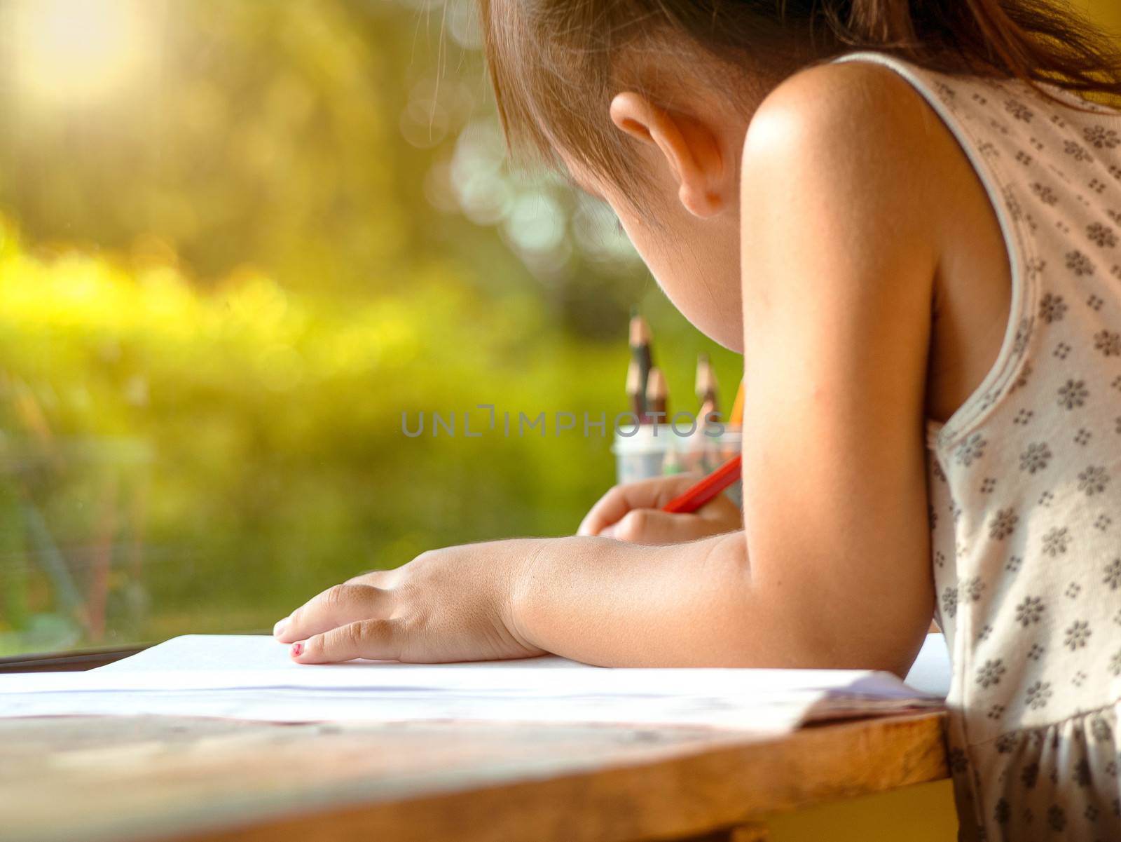 Cute Asian little girl using colored pencils to paint on paper, sitting at a table by the window in the house. Preschoolers study at home during the corona virus epidemic. by TEERASAK