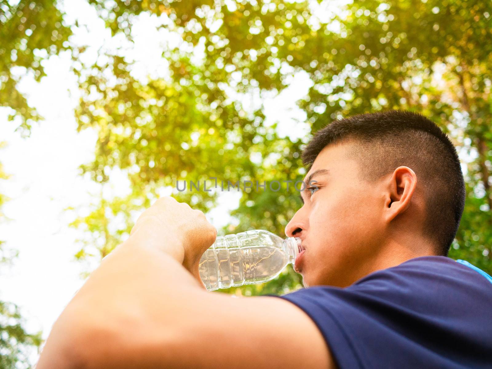 Asian teenage boy drinking water from bottle after jogging in the park. by TEERASAK