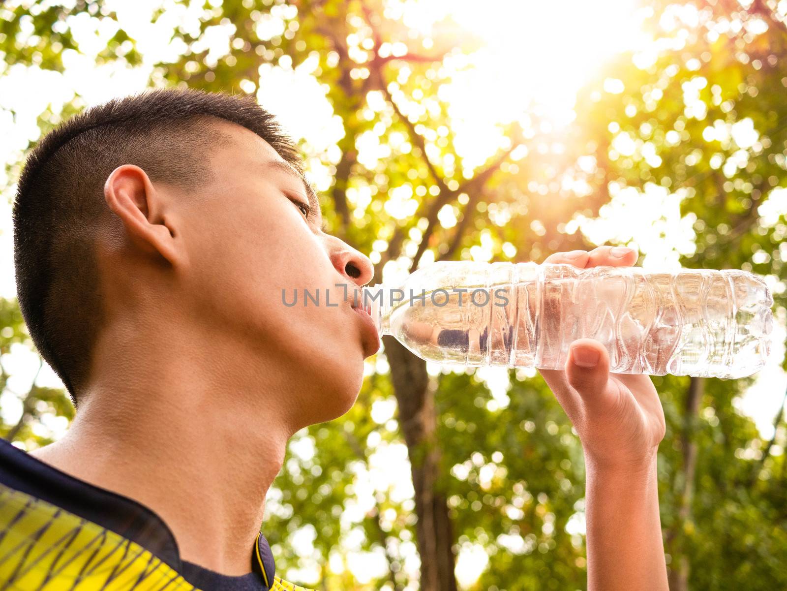 Asian teenage boy drinking water from bottle after jogging in the park. by TEERASAK