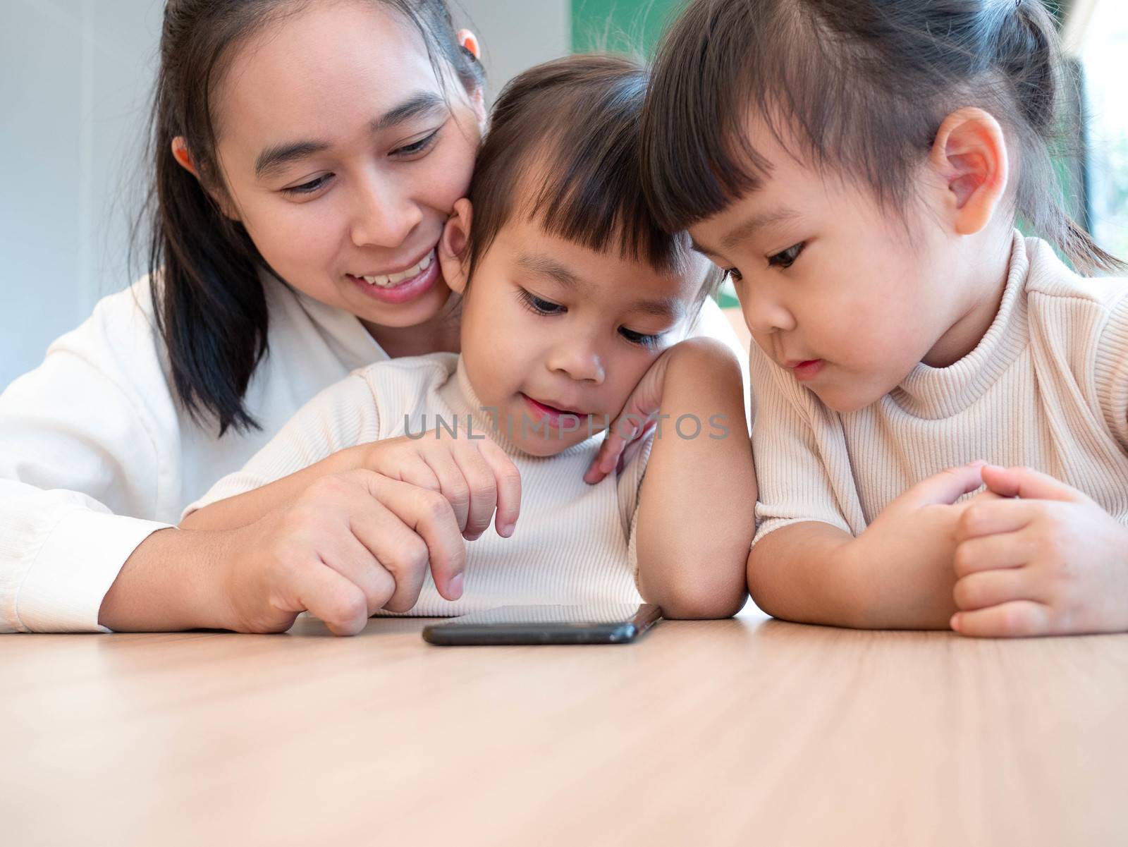 Asian beautiful young mother and her little daughters are enjoying playing games or entertaining using mobile apps on phone at home.