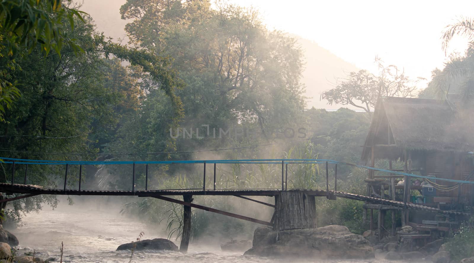 The beautiful view of fresh river stream water slowly flowing through natural rock beach with vaporizing in early morning and bamboo bridge over the river.