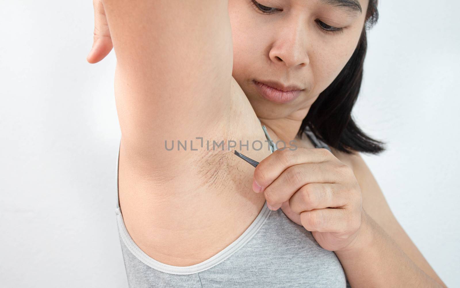 Close up of young woman plucking armpit underarm with tweezers isolated on grey background. Concept of Hygiene skin body care and beauty.