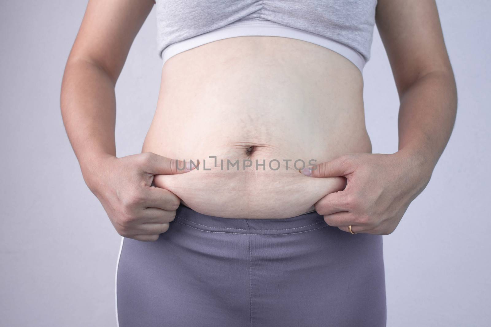 Close-up of woman's hand pinching excessive belly fat isolated on gray background. Woman fat belly. Obesity and Overweight Concept.