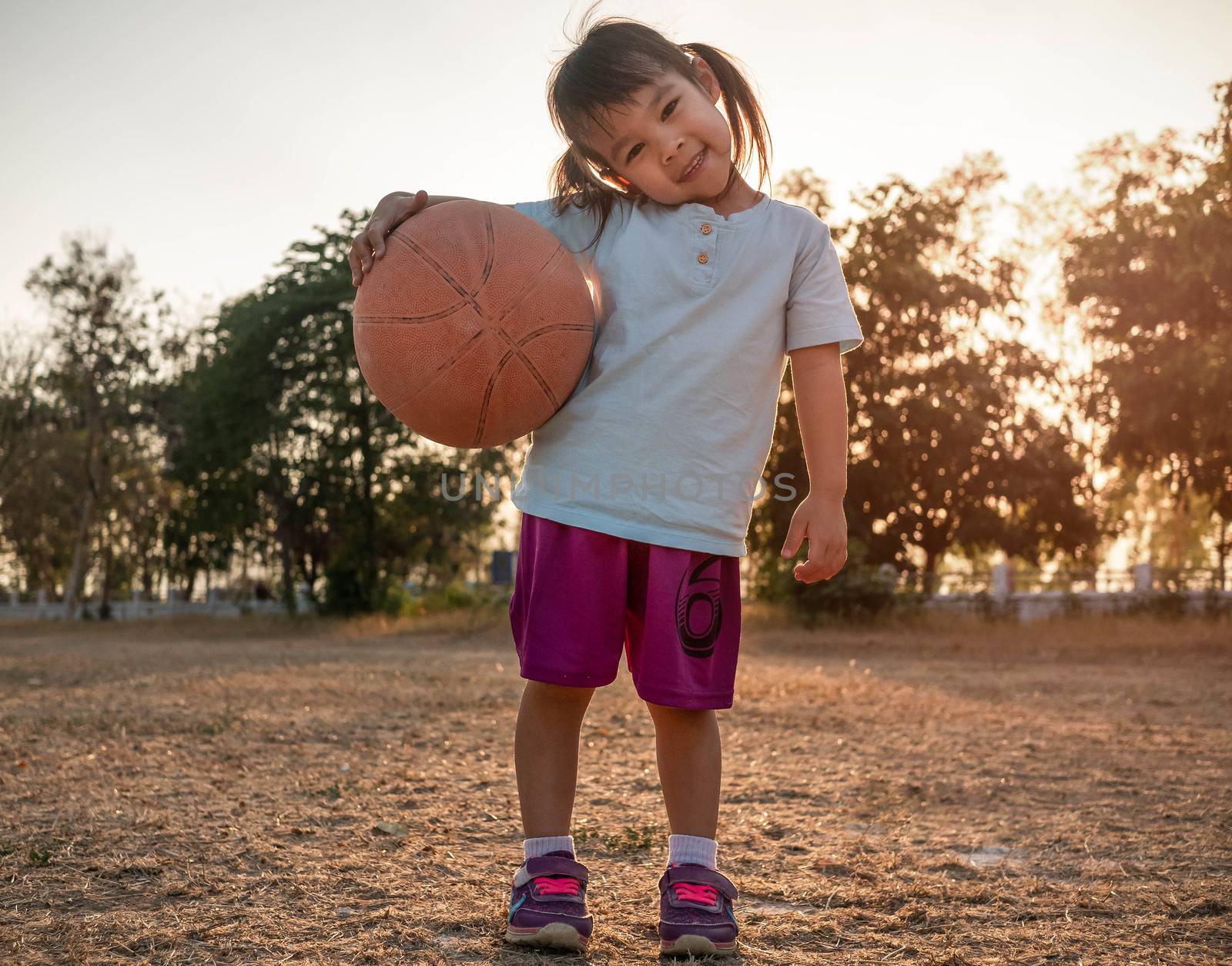 Cute Asian little girl in sportswear holding a basketball and looking at the camera at summer park. Healthy outdoor sport for young child. by TEERASAK