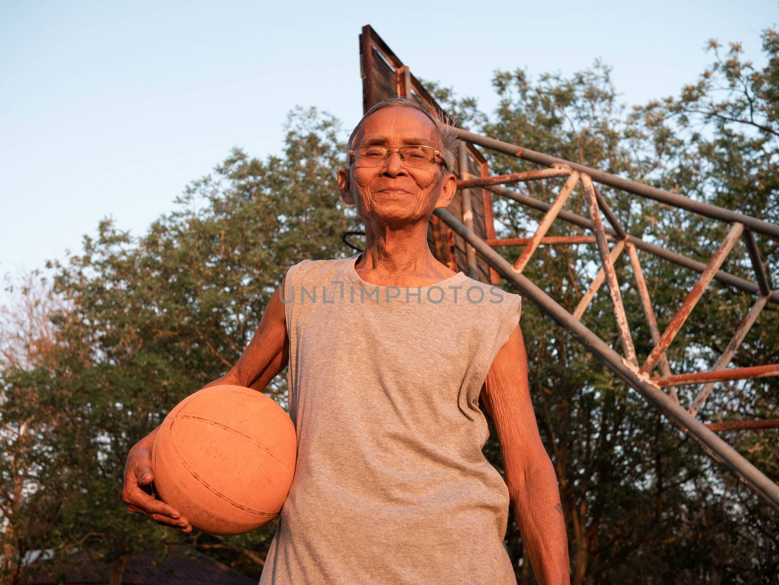 Asian elderly men in sportswear holding a basketball and looking at the camera at an outdoor basketball court on summer day. Healthy lifestyle and Healthcare concept. by TEERASAK