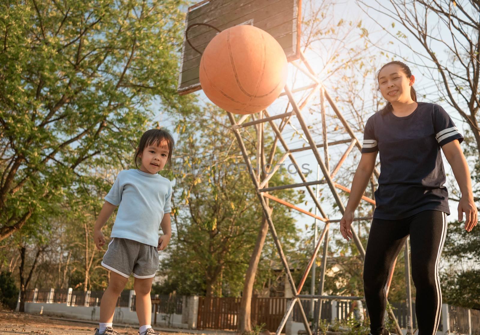 Cute little Asian child playing basketball with her mother on playground on summer day. Happy family spending free time together. by TEERASAK