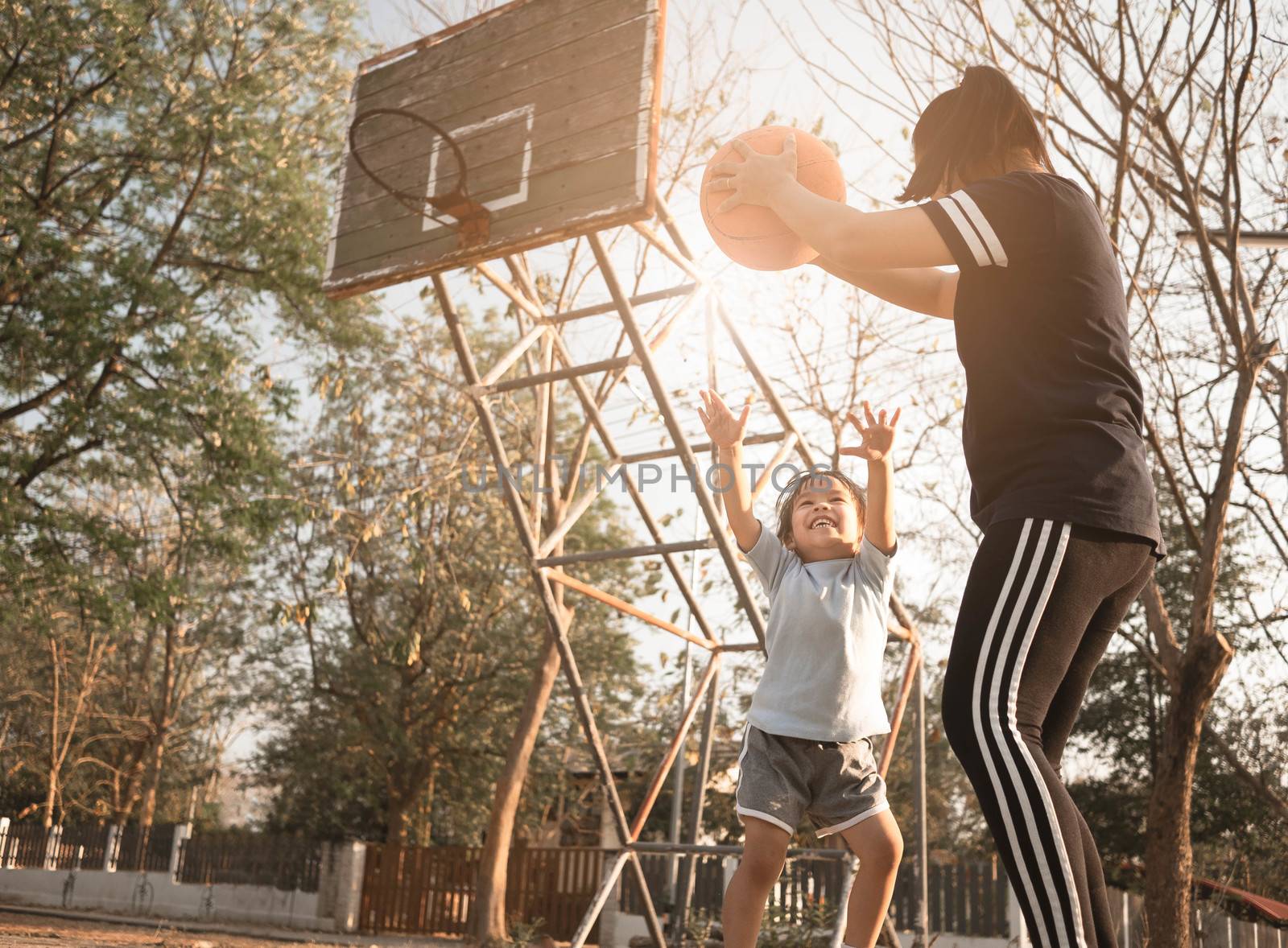 Cute little Asian child playing basketball with her mother on playground on summer day. Happy family spending free time together. by TEERASAK