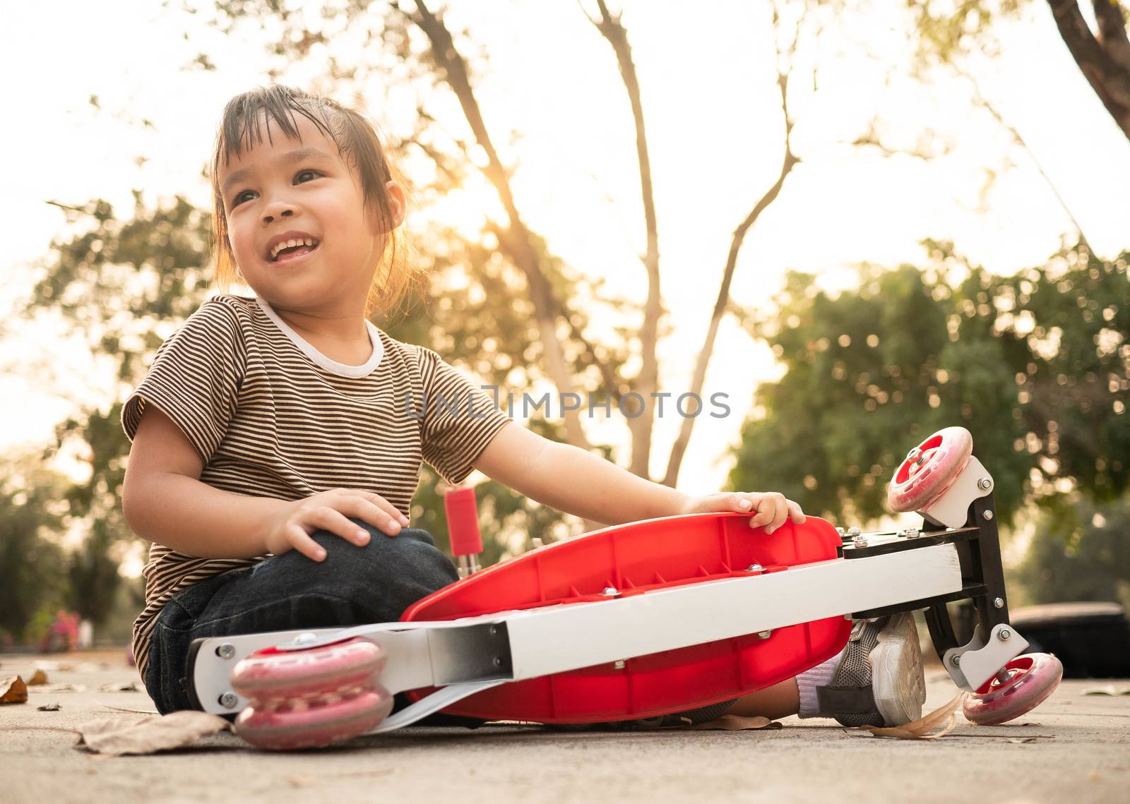 Cute Asian little girl sitting on the ground after falling off her scooter at summer park. Healthy outdoor sport for young child. by TEERASAK