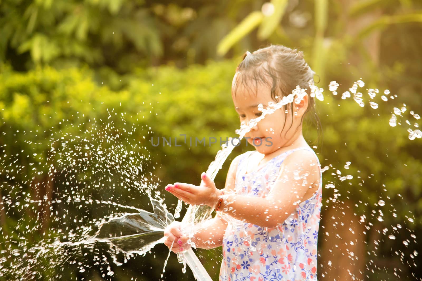 Cute little girl playing a rubber hose spraying water with sunlight in the backyard. Children enjoy outdoor activities on hot summer days.