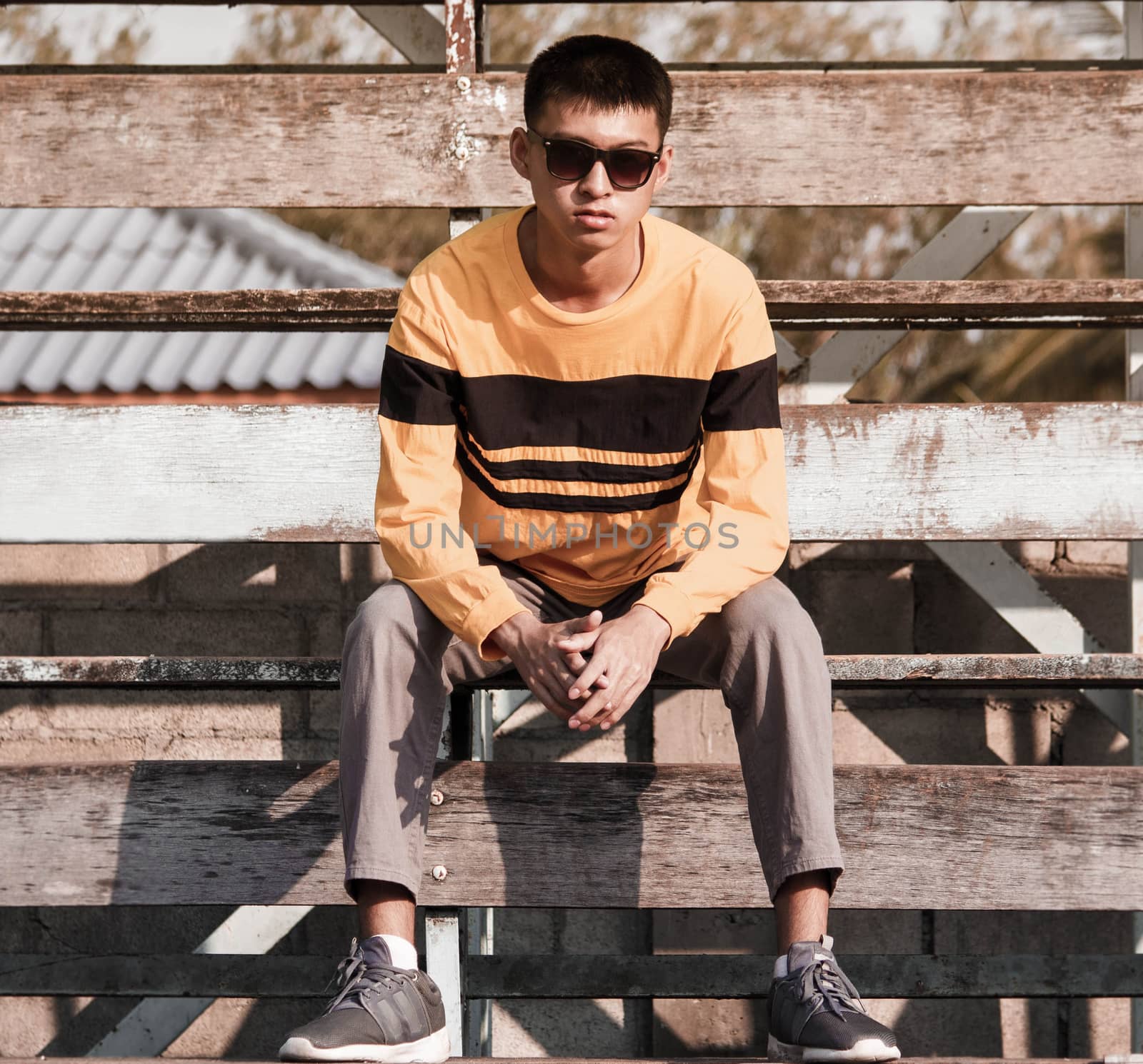 Portrait of a young handsome teenager boy in yellow long sleeve t-shirt and wear black glasses, sit on wooden stage outdoors. lifestyle people concept. by TEERASAK