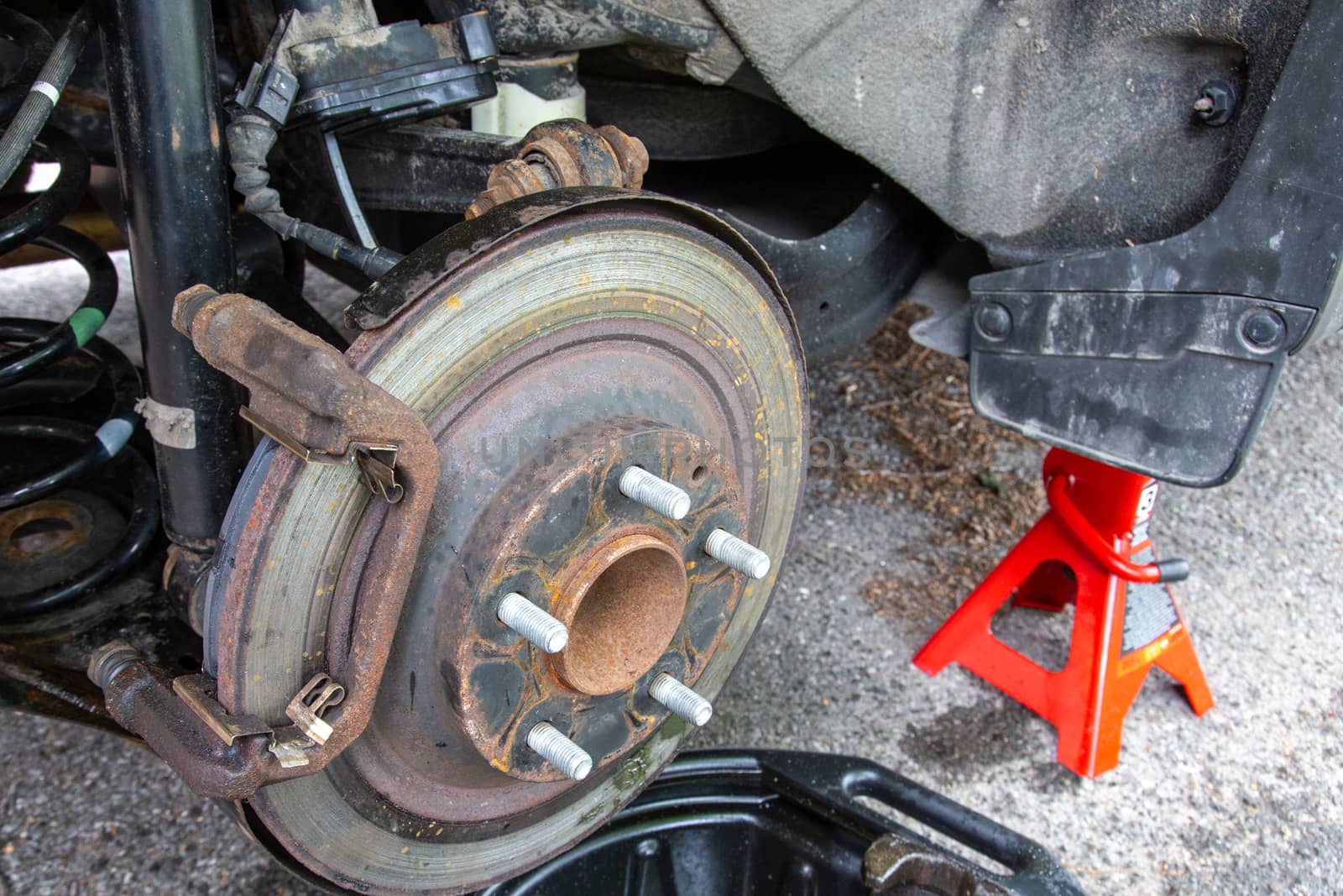 Brake work is being done on a car in a driveway. The vehicle hoisted up on a red jack with an exposed rear wheel, rusted rotor and removed brake caliper.