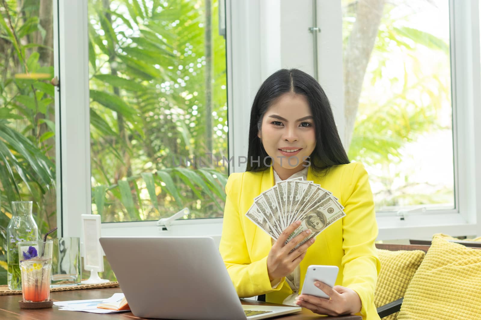 portrait of cheerful young Asian woman holding money banknotes and mobile phone with smile face in front of computer notebook at workplace