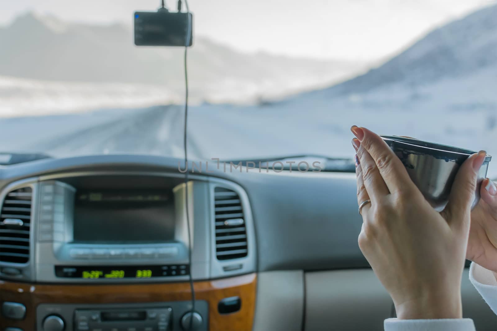 the hand of the girl in the passenger seat of the car holding hot tea from the mug from the thermos, view of the mountain winter road outside the car window