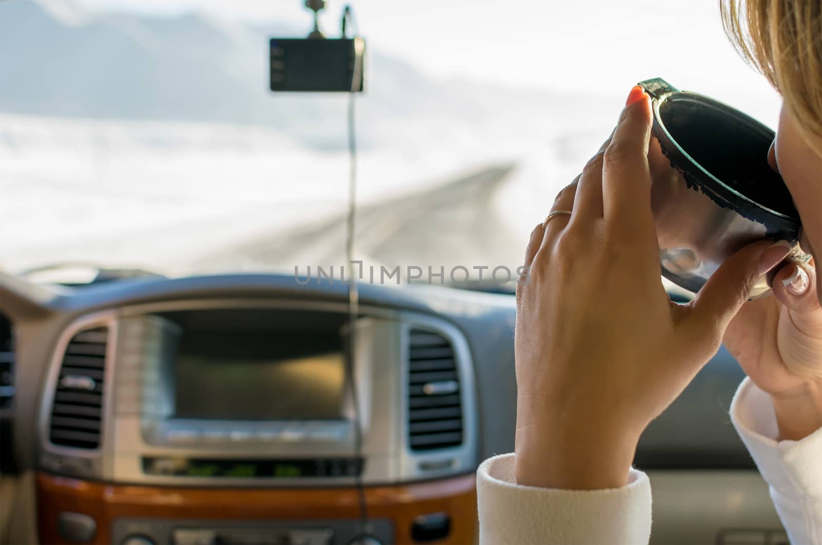 The girl in the passenger seat of the car drinking hot tea from a mug of a thermos, looking on the mountain winter road outside the car window by jk3030