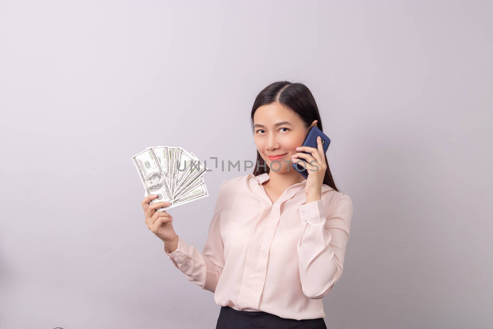 Asian beautiful woman holding banknote money in hand and mobile phone in another hand isolated on grey background. commercial business by phone concept by asiandelight