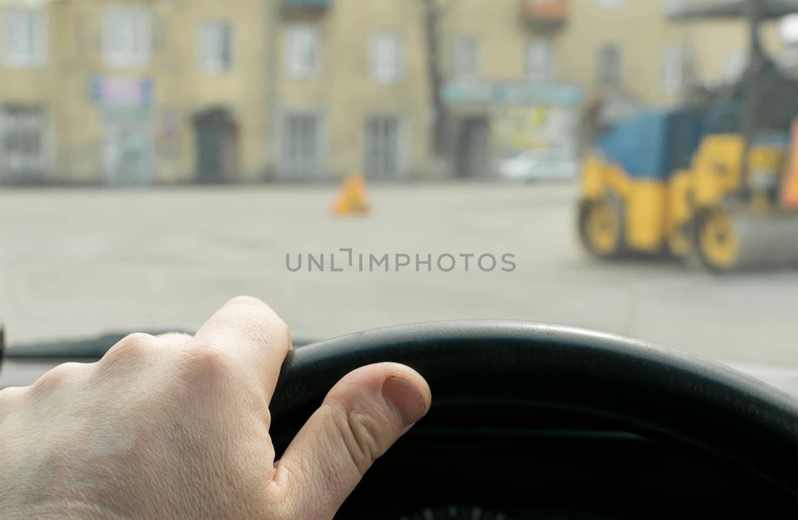 Close-up, the driver's hand on the steering wheel of the car on the background of construction road machinery, road paver and road repair in cloudy weather
