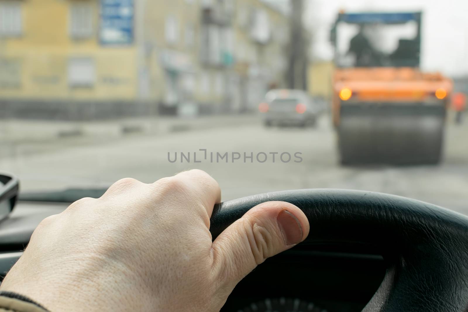 Close-up, the driver's hand on the steering wheel of the car on the background of construction road machinery, road paver and road repair in cloudy weather