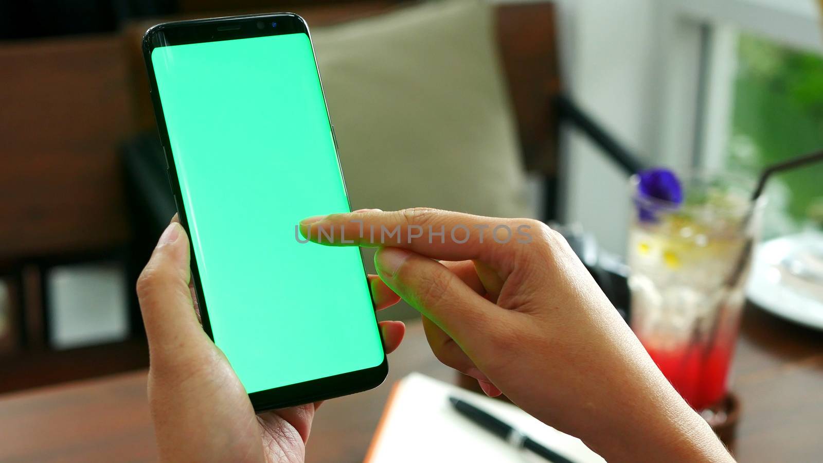 woman hand holding mobile smart phone with blank green screen, use finger slide on green screen at cafe shop with drink at background