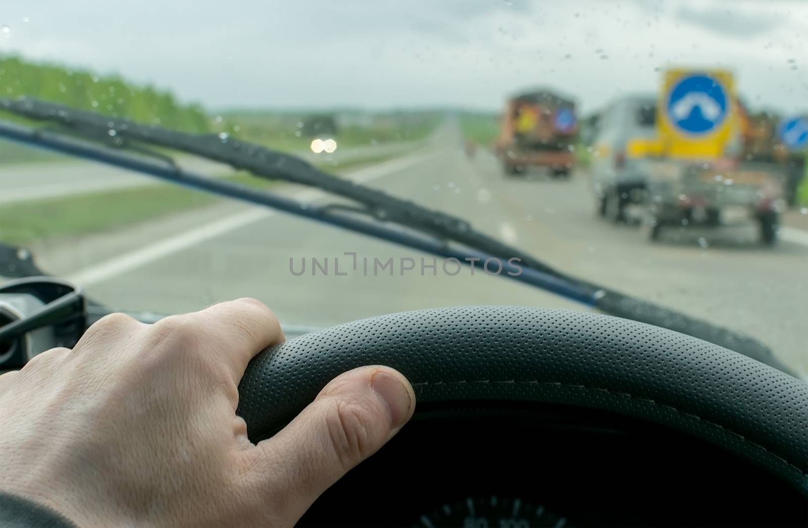 The hand of the driver who moves on a country road in rainy cloudy weather background drops of rain on a windshield and special equipment for repair of the road with warning signs
