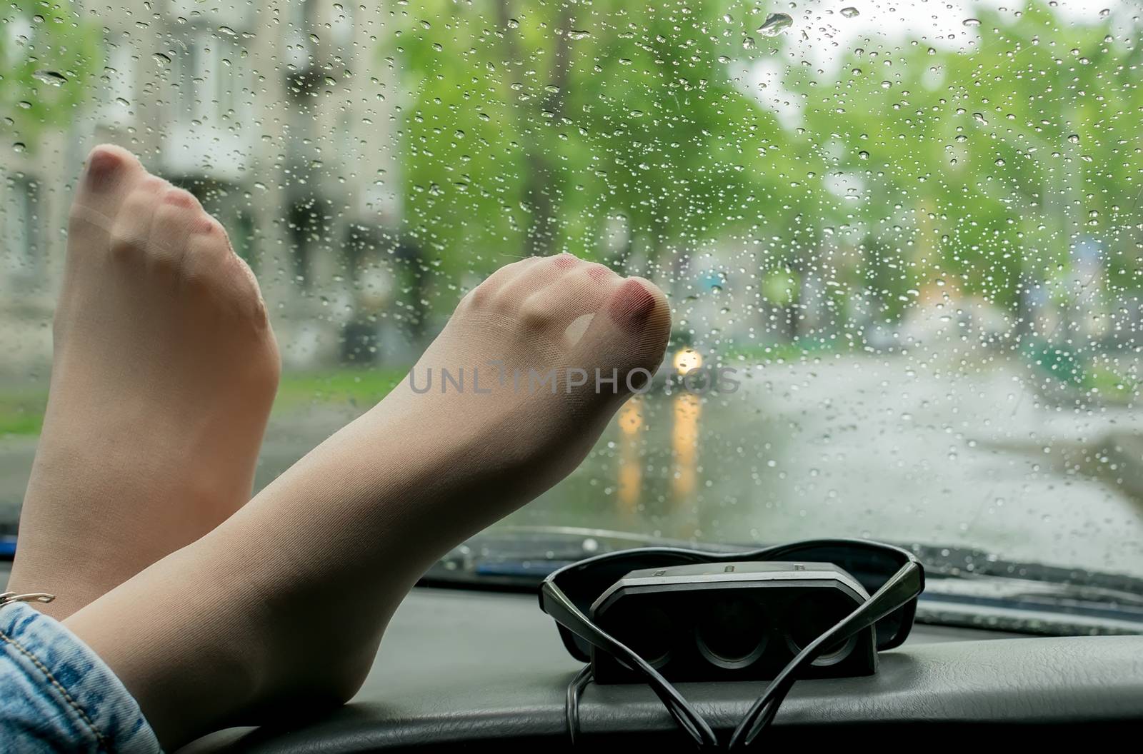 female feet in jeans, tights and with painted nails lie on the panel of the car against the wet windshield on the background of the street